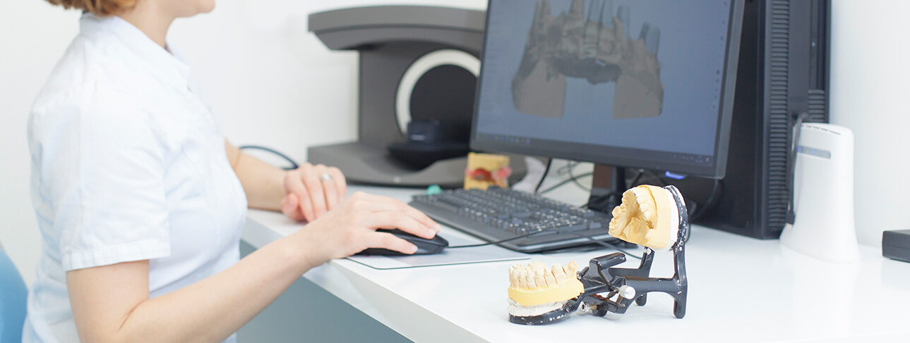 woman working on a computer with an image of teeth on it, while a model set of teeth are next to her as well