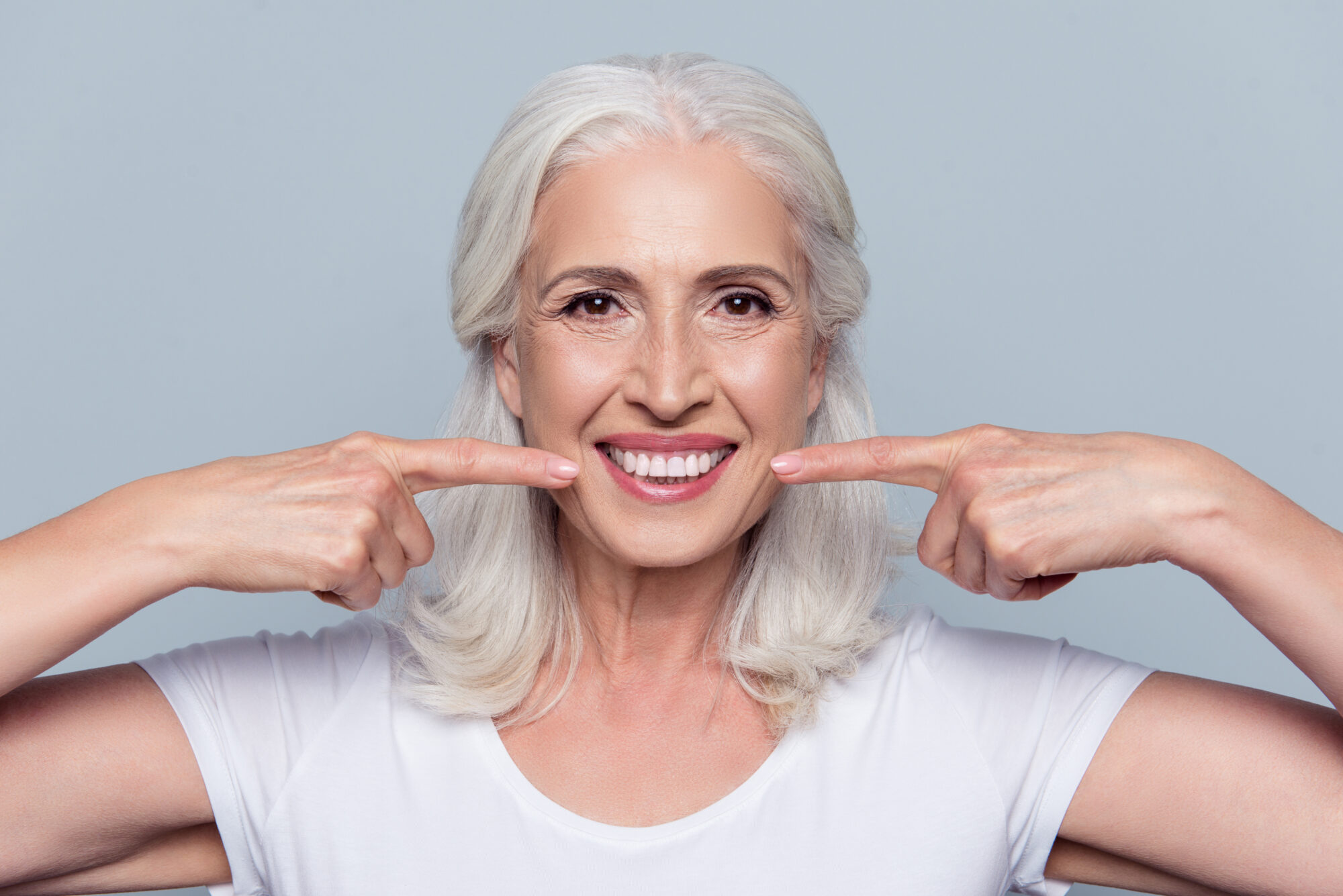 Close up portrait of happy with beaming smile female pensioner pointing on her perfect clear white teeth, isolated on gray background