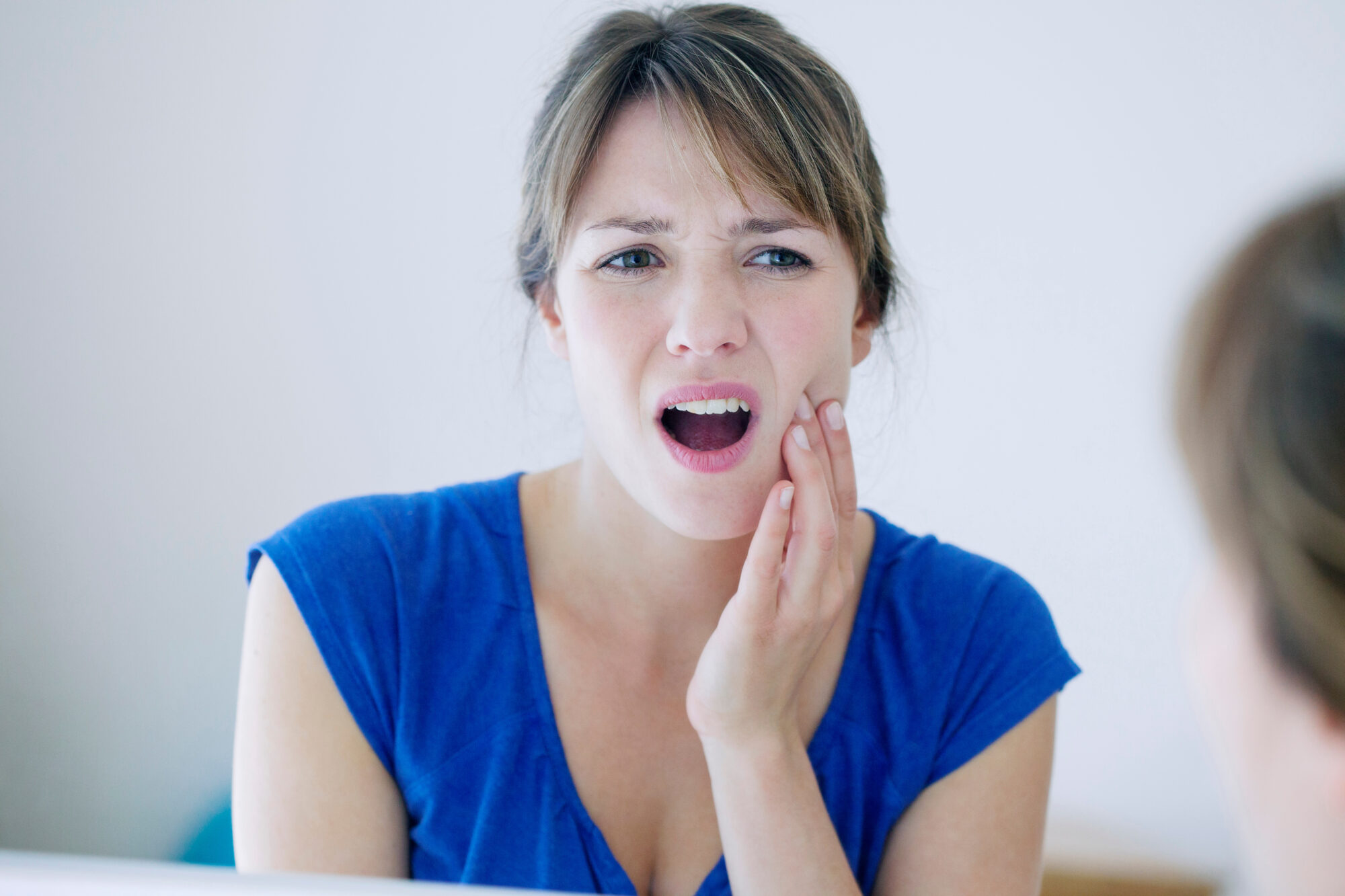 Photograph of a woman. Her face shows that she is in pain, and she has her hand pressed to the side of her face where the jaw meets the skull, or at the TMJ joint.